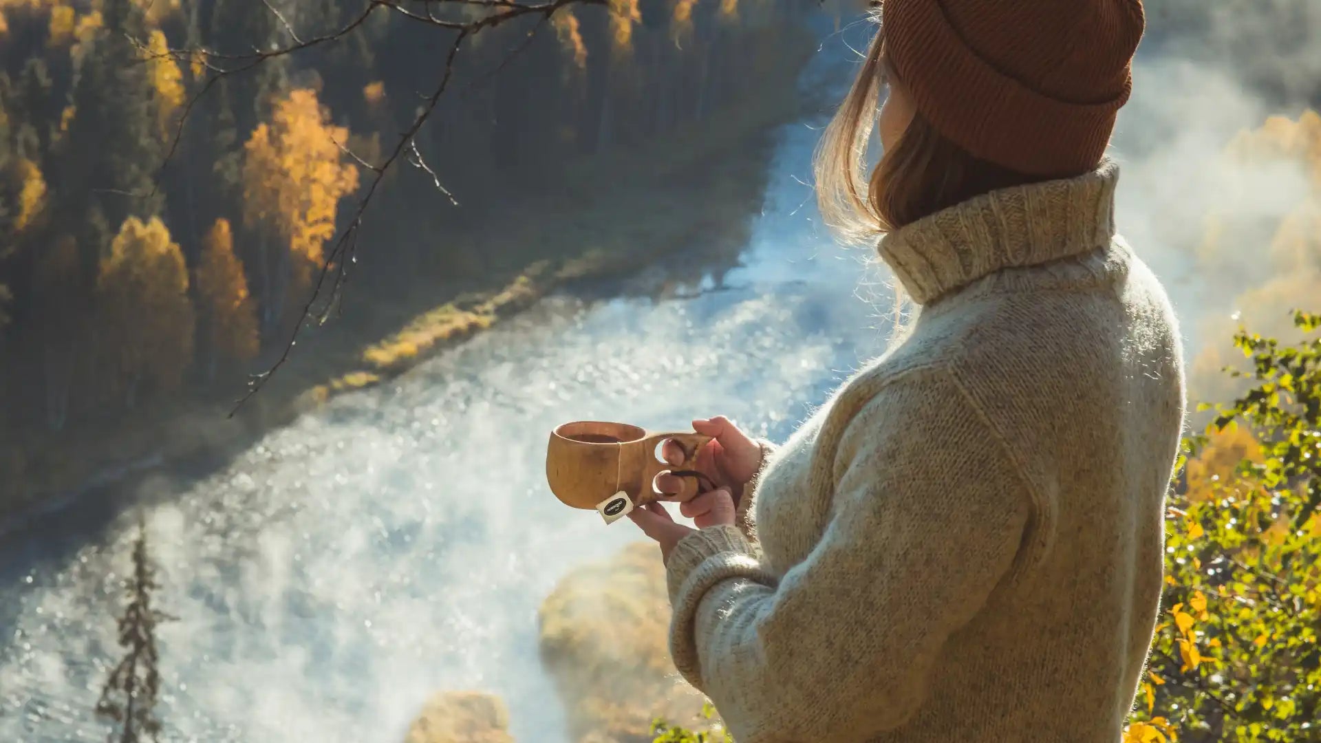 Ihminen lämpimässä villapaidassa ja hatussa, joka pitää puista kupia, katsellen maisemaa, jossa on vesiputous ja syksyn puita.